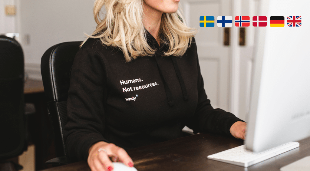Blond kvinna som sitter framför en stationär dator med en hoodie från Wndy. Svenska, norska, danska, tyska och uk flaggor i hörnet av bilden